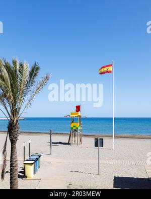 Strand mit einer Palme im Vordergrund. Klarer blauer Himmel, Tiefsee, Ozean, winkende Flagge Spaniens, Sand, farbige Mülltonnen, Rettungsstand, Sommer Stockfoto