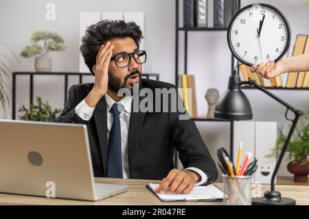 Indischer Geschäftsmann, der an einem Büro-Laptop arbeitet, mit Angst, die Zeit auf Uhr zu überprüfen, zu spät zur Arbeit zu kommen, in Verspätung zu sein, Deadline. Der Manager-Freiberufler schaut auf die Stunde, Minuten, um pünktlich zu sein Stockfoto