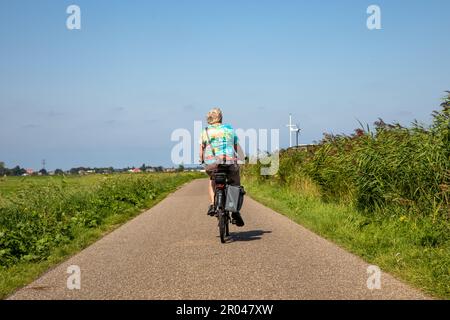 Ältere Frau mit Kameratasche, die auf einem Elektrofahrrad im südholländischen Dorf Sassenheim in den Niederlanden fährt. An einem blauen Tag. Stockfoto