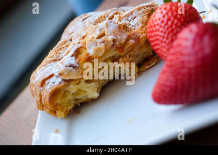 Ein gebissenes Croissant mit Mandelflocken und Puderzucker auf weißem Teller zwei große rote Erdbeeren Foto schräg Platz für Textwerbung köstlich Stockfoto