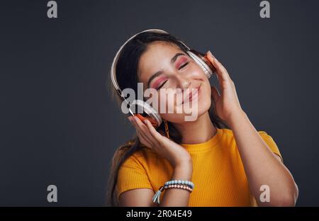 Dieses Lied gibt mir ein gutes Gefühl. Ein attraktives Teenager-Mädchen, das allein vor einem dunklen Hintergrund steht und Musik über Kopfhörer hört. Stockfoto