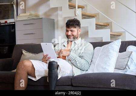 Glücklicher Mann mit behindertengerechter Beinprothese, der zu Hause auf der Couch sitzt und ein Tablet benutzt. Stockfoto