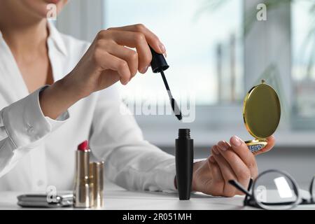 Junge Frau, die einen kosmetischen Taschenspiegel und Mascara an einem weißen Tisch im Haus benutzt, Nahaufnahme Stockfoto