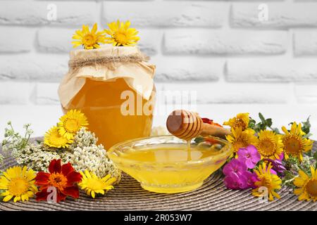 Köstlicher Honig und Dipper mit verschiedenen Blumen auf Korbmatte nahe der weißen Backsteinwand Stockfoto