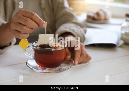 Eine Frau, die den Teebeutel aus dem Becher am weißen Holztisch im Haus nimmt, Nahaufnahme. Platz für Text Stockfoto