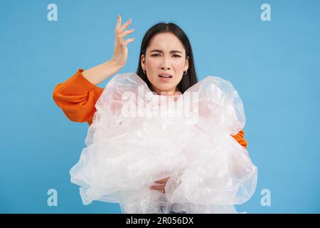 Genervte asiatische Freiwillige, die Plastikabfall in der Hand hält, mit enttäuschtem Gesicht schreit, die Verschmutzung hasst, steht über blauem Hintergrund. Stockfoto