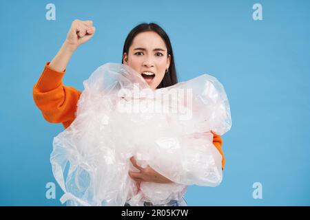 Wütender Öko-Aktivist, der Plastikabfälle hält, die Faust erhebt und für die Umwelt kämpft, Müll sortiert, recycelt, blauer Hintergrund Stockfoto