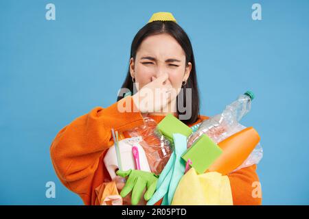 Das Porträt einer Frau hält ihre Nase vor der Nase, hält stinkenden Müll, schmutzige Plastikgegenstände und leere Flaschen mit schlechtem Geruch, steht über blau Stockfoto