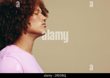 Ein junger Mann, der in einem Studio mit geschlossenen Augen steht, umarmt seine Sommersprossen-Haut und Ingwer-Afro-Haare. Ein junger und gutaussehender Mann, der Selbstvertrauen ausdrückt Stockfoto