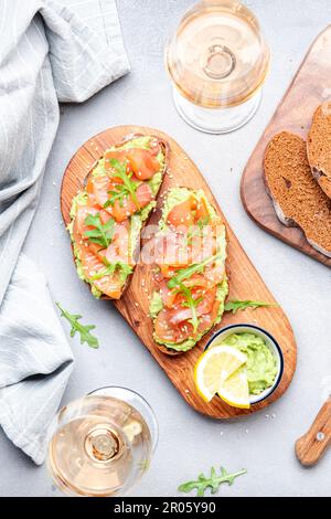 Gourmet-Mittagessen mit Avocado-Lachs-Toasts auf Roggenbrot und Rosenweingläsern, grauer Tischhintergrund, Blick von oben Stockfoto