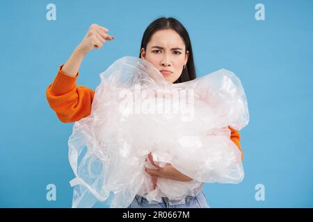 Wütender Öko-Aktivist, der Plastikabfälle hält, die Faust erhebt und für die Umwelt kämpft, Müll sortiert, recycelt, blauer Hintergrund Stockfoto