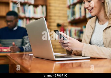 Vielleicht hilft mir eine Lerngruppe. Eine Universitätsstudentin, die ihr Handy benutzt, während sie hinter einem Laptop sitzt. Stockfoto