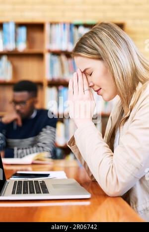 Sie betet, dass sie diese Tests besteht. Eine junge Frau, die gestresst aussieht, während sie hinter einem Laptop sitzt. Stockfoto