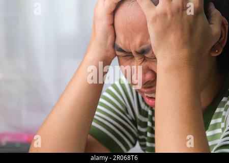 Junge Frau mit Kopfschmerzen vor weißem Vorhang im Zimmer. Eine Erwachsene Frau, die wegen Kopfschmerzen oder Migräne den Kopf berührt. Hände auf den Kopf. Stockfoto