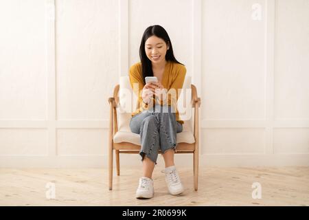 Fröhliche asiatische Frau, die im Sessel sitzt und Handy benutzt Stockfoto