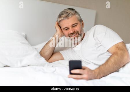 Fröhlicher, grauhaariger europäer, der im Bett lag und telefonierte Stockfoto