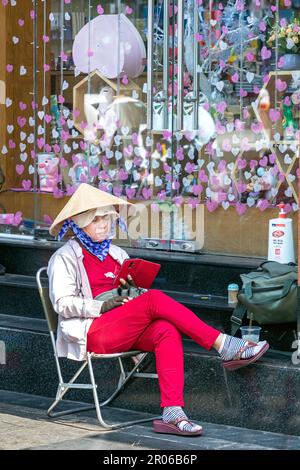 Vietnamesische Straßenverkäuferin trägt Bambushut und benutzt ihr Handy im Stadtzentrum von Ho Chi Minh City, Vietnam Stockfoto