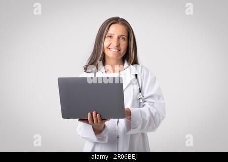 Medizinische Online-Forschung. Eine leitende Ärztin, die einen Laptop in der Hand hält, vor der Kamera lächelt und über einem hellen Hintergrund steht Stockfoto