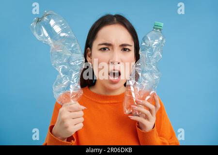 Wütende asiatische Frau mit zwei Plastikflaschen mit Stirnrunzeln, Mädchen mit umweltfreundlichem Lebensstil, Drang, Abfall zu recyceln, blauer Hintergrund Stockfoto