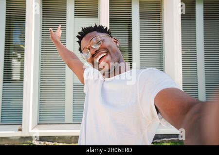 Lächelnder junger afroamerikanischer Student, der in die Kamera schaut und ein Selfie macht, mit seinem Handy draußen. Porträt eines fröhlichen Jahrtausendsmannes mit einem zahnigen Lächeln. Hochwertiges Foto Stockfoto