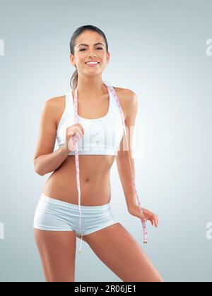 Nichts fühlt sich so gut an wie fit. Studiofoto einer jungen Frau, die mit einem Maßband vor grauem Hintergrund posiert. Stockfoto