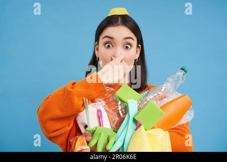 Das Porträt einer Frau hält ihre Nase vor der Nase, hält stinkenden Müll, schmutzige Plastikgegenstände und leere Flaschen mit schlechtem Geruch, steht über blau Stockfoto