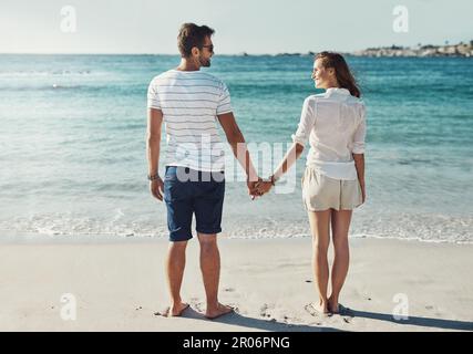 In den Sonnenuntergängen zusammenhalten... das ist wahre Liebe. Rückblick auf ein junges Paar, das im Sommer den Sonnenuntergang an der Küste beobachtet. Stockfoto
