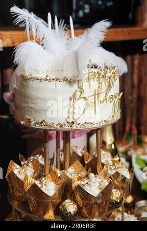 Geburtstagskuchen mit Federn. goldverzierte Cupcakes. Geburtstagsbonbons auf einem Tisch. Vertikale Aufnahme von Partygeschenken Stockfoto