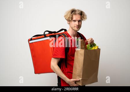 Ein attraktiver Zusteller, ein Kurier, der Lebensmittel und Getränke aus dem Laden mitbrachte, ein Supermarkt, isoliert auf weißem Studiohintergrund. Stockfoto