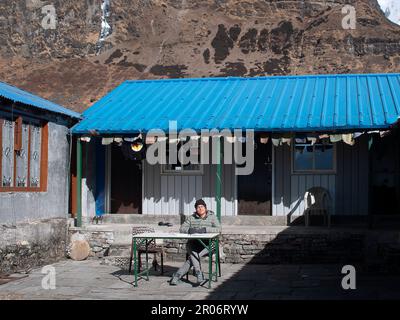 Eine weibliche Rucksacktouristin sieht melancholisch aus im Machhapuchhare Basislager (Nepal) und malt in ihrem Tagebuch mit Wasserfarben und Gästehaus im Hintergrund. Stockfoto