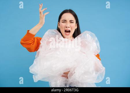 Genervte asiatische Freiwillige, die Plastikabfall in der Hand hält, mit enttäuschtem Gesicht schreit, die Verschmutzung hasst, steht über blauem Hintergrund Stockfoto