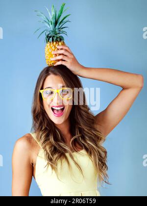 Was ist das Leben ohne ein wenig Dummheit? Studioaufnahme einer jungen Frau, die eine Ananas auf dem Kopf balanciert, vor einem blauen Hintergrund. Stockfoto