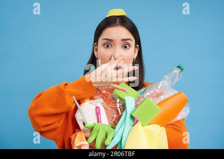 Das Porträt einer Frau hält ihre Nase vor der Nase, hält stinkenden Müll, schmutzige Plastikgegenstände und leere Flaschen mit schlechtem Geruch, steht über blau Stockfoto