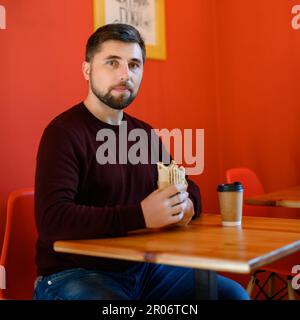 Ein Mann mit Bart sitzt in einem Fast-Food-Restaurant und hält ein Schawarma in der Hand. Er hat ein Glas Kaffee, eine rote Wand und einen Holztisch. Stockfoto