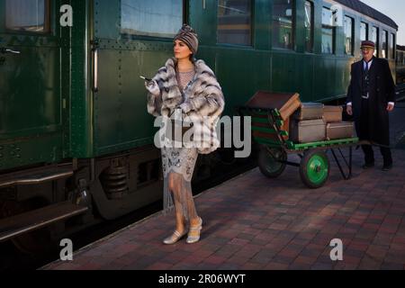 Nachstellungsszene auf dem Bahnsteig in der Nähe eines authentischen 1927 First Class Zugwaggons, wo eine schicke 1920er Dame auf ihr Gepäck wartet Stockfoto