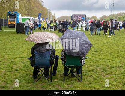 Im strömenden Regen sitzen ein Paar auf faltbaren Stühlen unter Sonnenschirmen, um die historische Krönung von König Karl III. Auf einem großen Bildschirm in London zu beobachten Stockfoto