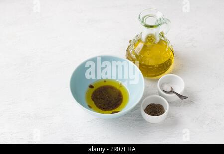 Eine blaue Schüssel mit Olivenöl, Gewürzen und Balsamsauce, umgeben von einer Flasche Olivenöl, einem Salz- und Pfefferstreuer auf hellgrauem Hintergrund. Tradit Stockfoto