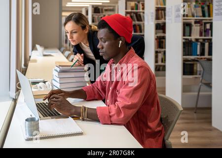 Interessierte afroamerikanische Schüler, die auf dem Laptop tippen, sind in der Nähe fokussierter College-Lehrer, die auf den Bildschirm schauen Stockfoto