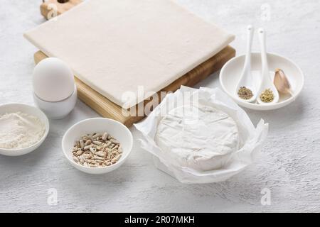 Zutaten für einen köstlichen hausgemachten Kuchen oder Snack: Blätterteig, weißer Schimmelkäse (Camembert oder Brie), Ei, Gewürze, Samenmischung und Mehl auf einem Light Stockfoto