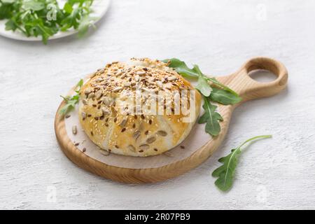 In Blätterteigkäse gebacken, bestreut mit Samen, dekoriert mit frischen Rucola-Blättern auf einem Holzbrett auf grauem, strukturiertem Hintergrund Stockfoto