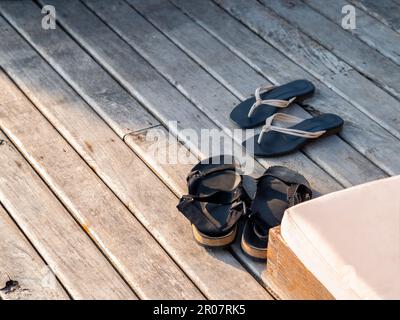 Leere schwarze Sandalen von Männern und Frauen mit Sandalen wurden auf der hölzernen Terrasse ausgezogen und gingen bei Sonnenuntergang am Strand spazieren. Sommerferien, Stockfoto