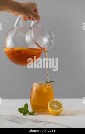 Eine Frau gießt Eistee mit Zitrone aus einer Kanne in ein Glas. Sommergetränke. Stockfoto