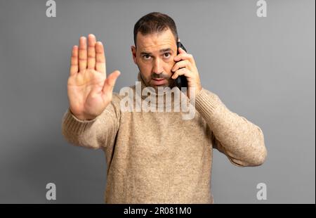 Ein bärtiger Latino in seinen 40s Jahren, der einen beigen Rollkragenpulli trägt und mit einem ernsten Gesichtsausdruck auf dem Handy spricht, während er seine Hand in einem Stoppschild hebt Stockfoto