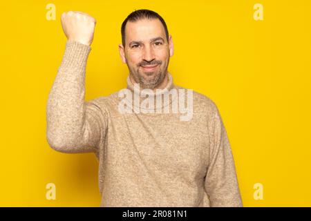 Bärtiger hispanischer Mann in seinen 40s Jahren mit beigefarbenem Rollkragenpullover, der seine Faust erhob und mit einem siegreichen Gesichtsausdruck lächelte, isoliert über einem gelben Studio Stockfoto