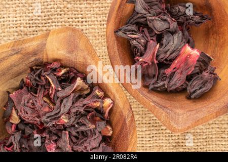 Trockener, duftender Karkade-Tee in zwei Holzbechern auf einem Jutetuch, Makro, Draufsicht. Stockfoto