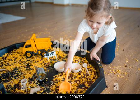 Kleines Mädchen, das mit sensorischen Flocken im Sandkasten spielt. Sensorische Entwicklung und Erfahrungen, Themenaktivitäten mit Kindern, Feinmotorik Stockfoto