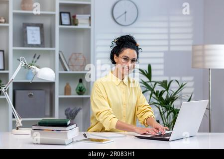 Porträt einer glücklichen, erfolgreichen Frau bei der Arbeit im Home Office, einer hispanischen Frau, die lächelt und auf die Kamera schaut, am Tisch im Wohnzimmer sitzt, mit einem Laptop aus der Ferne arbeitet, online lernt. Stockfoto