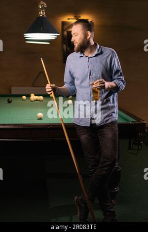 Ein bärtiger Mann, der Billard spielt, steht neben einem Tisch mit einem Stichwort in der Hand und hält ein Glas Bier und denkt über das Spiel nach Stockfoto