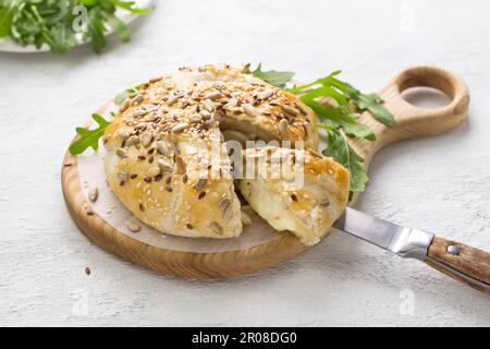 In Blätterteigkäse gebacken, bestreut mit Samen, dekoriert mit frischen Rucola-Blättern auf einem Holzbrett auf grauem, strukturiertem Hintergrund Stockfoto