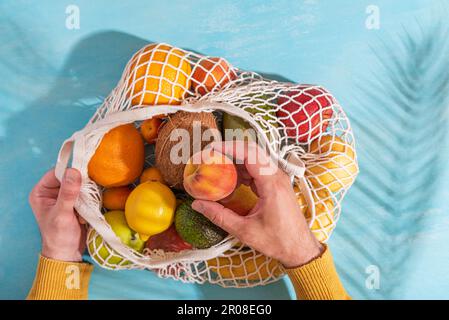 Männerhand, die Avocadofrüchte aus einer Einkaufstasche mit tropischen Früchten nimmt. Blauer Hintergrund mit Palmenblattschatten Stockfoto